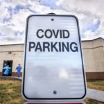 A sign says “COVID Parking” in front of a building, from which two people in personal protective equipment are walking.