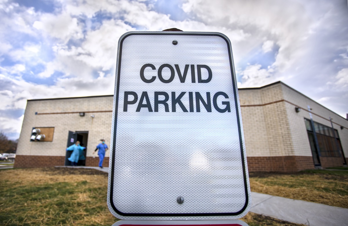 A sign says “COVID Parking” in front of a building, from which two people in personal protective equipment are walking.