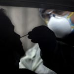 A health care worker with a N-95 mask and safety glasses is giving a nasal swab test to a man in a car.
