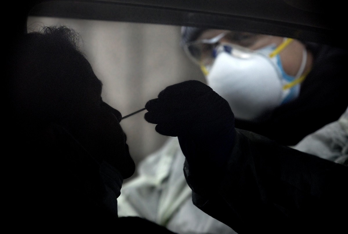 A health care worker with a N-95 mask and safety glasses is giving a nasal swab test to a man in a car.