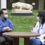 A man and woman sit outside at a wooden table with a statue of the Penn State Nittany Lion in the background. They both wear face masks and the woman is wearing glasses and hospital scrubs under a lab coat. The man’s arms are crossed.
