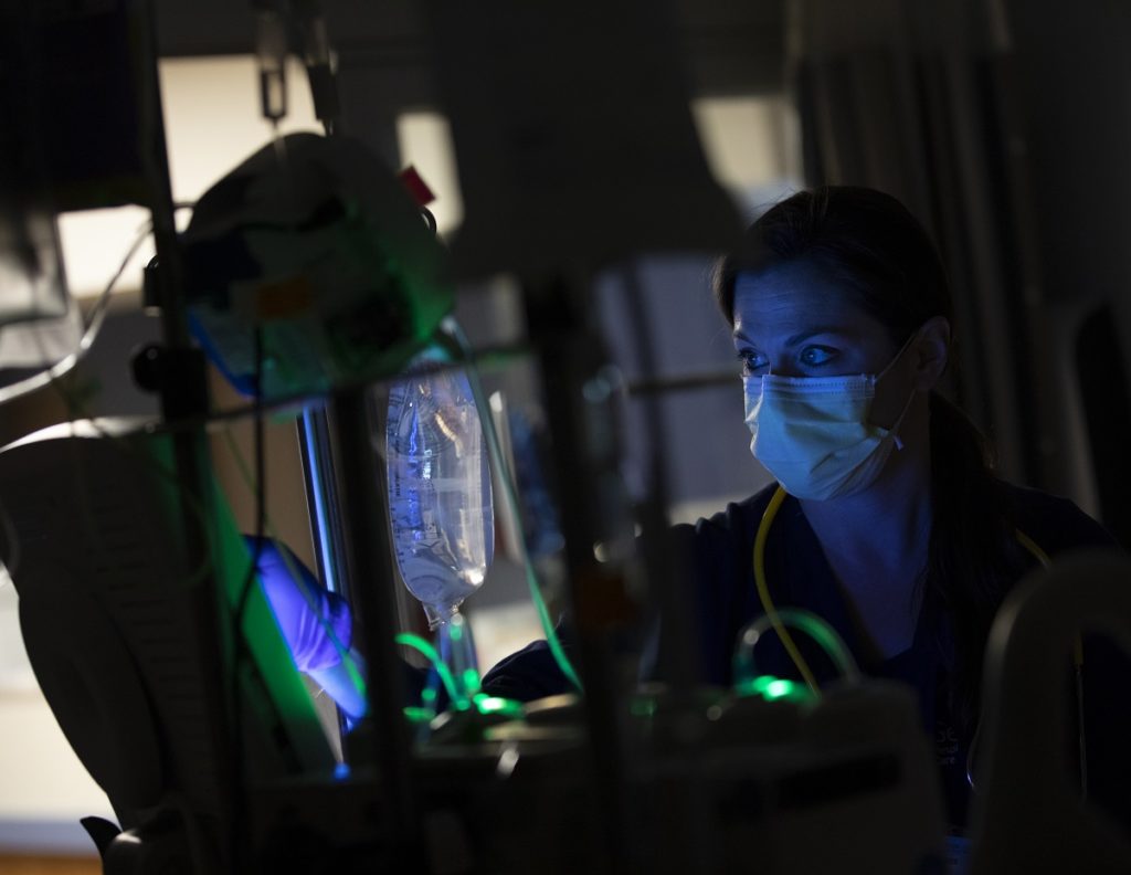 Erica Simpson, a respiratory therapist with Penn State Health Holy Spirit Medical Center, sits by a patient who is lying in bed. It is night, and a light on the patient reflects on Simpson’s face. A saline bag hangs between them. Simpson is wearing a face mask and scrubs. Part of the side of the patient’s face is visible. 