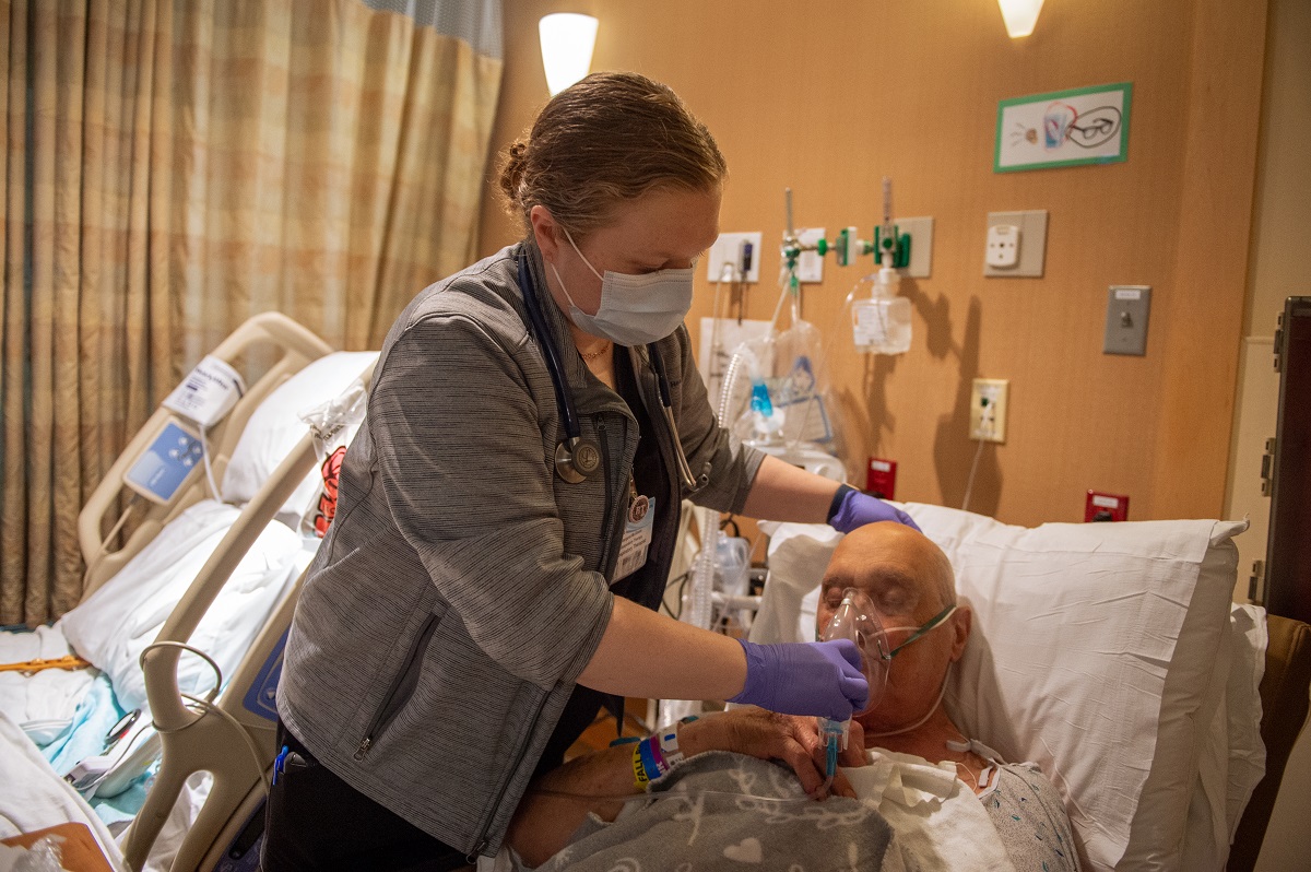 Margaret McGinnis, a respiratory therapist with Penn State Health St. Joseph, holds an oxygen mask to the face of a male patient who is lying in bed with his eyes closed. McGinnis is wearing scrubs, a sweat jacket, a lanyard with a nametag and a face mask. Behind her is another hospital bed.