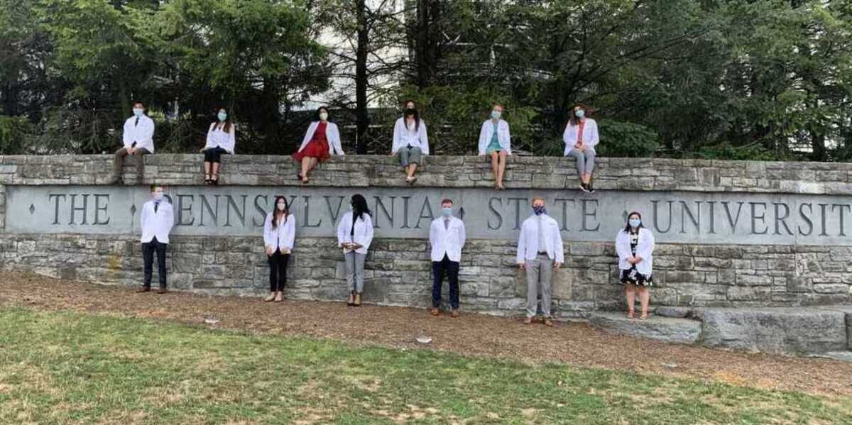Medical school students wearing white physician coats stand and sit around a gray, stone wall with the words “Penn State University” carved into it. A row of trees stand in the background.
