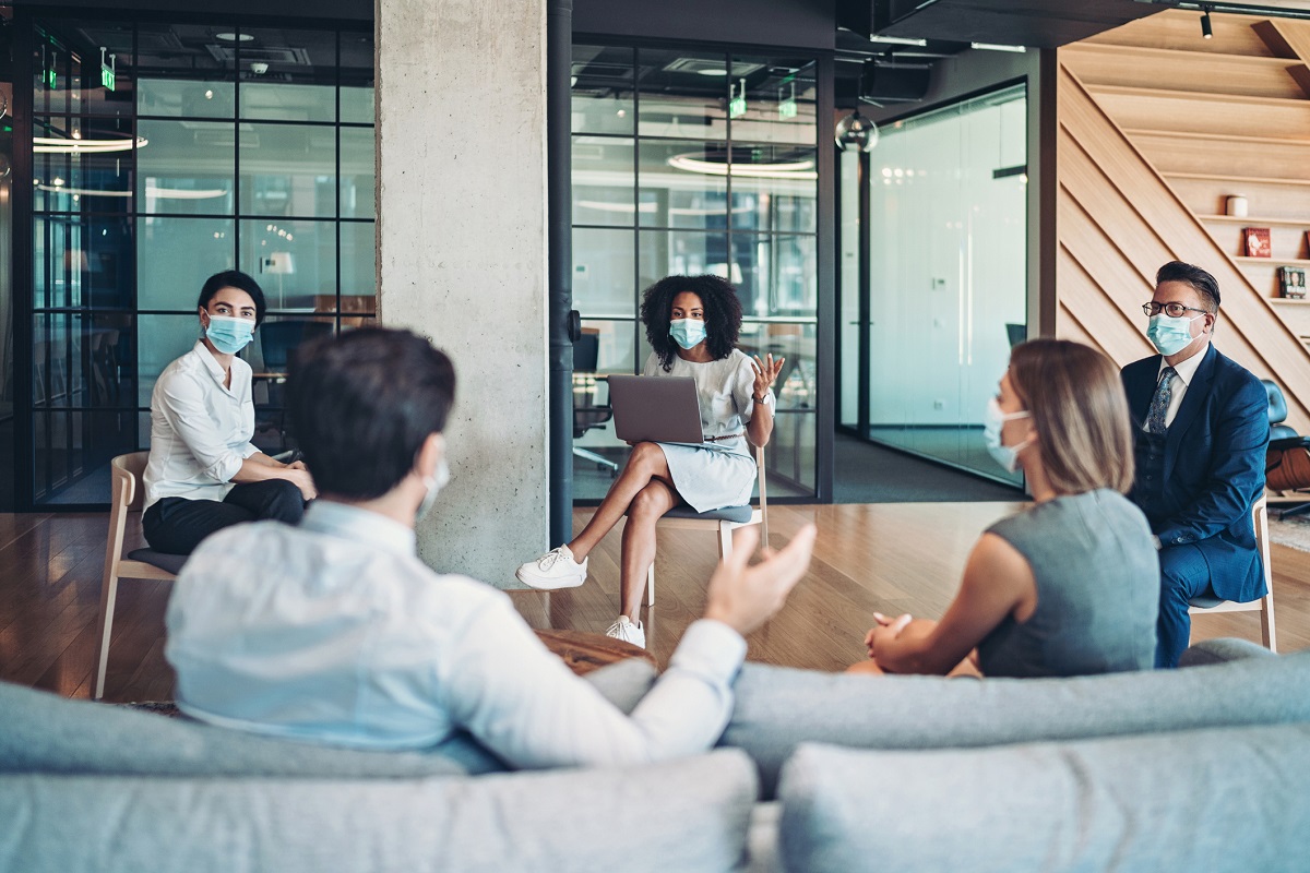 Five people in masks and business attire conduct a meeting in an office building.