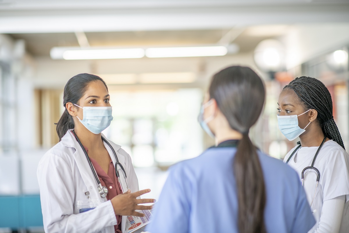 Three women with long hair pulled back in a ponytail and wearing face masks stand in a huddle. Two of them have a stethoscope wrapped around their necks.