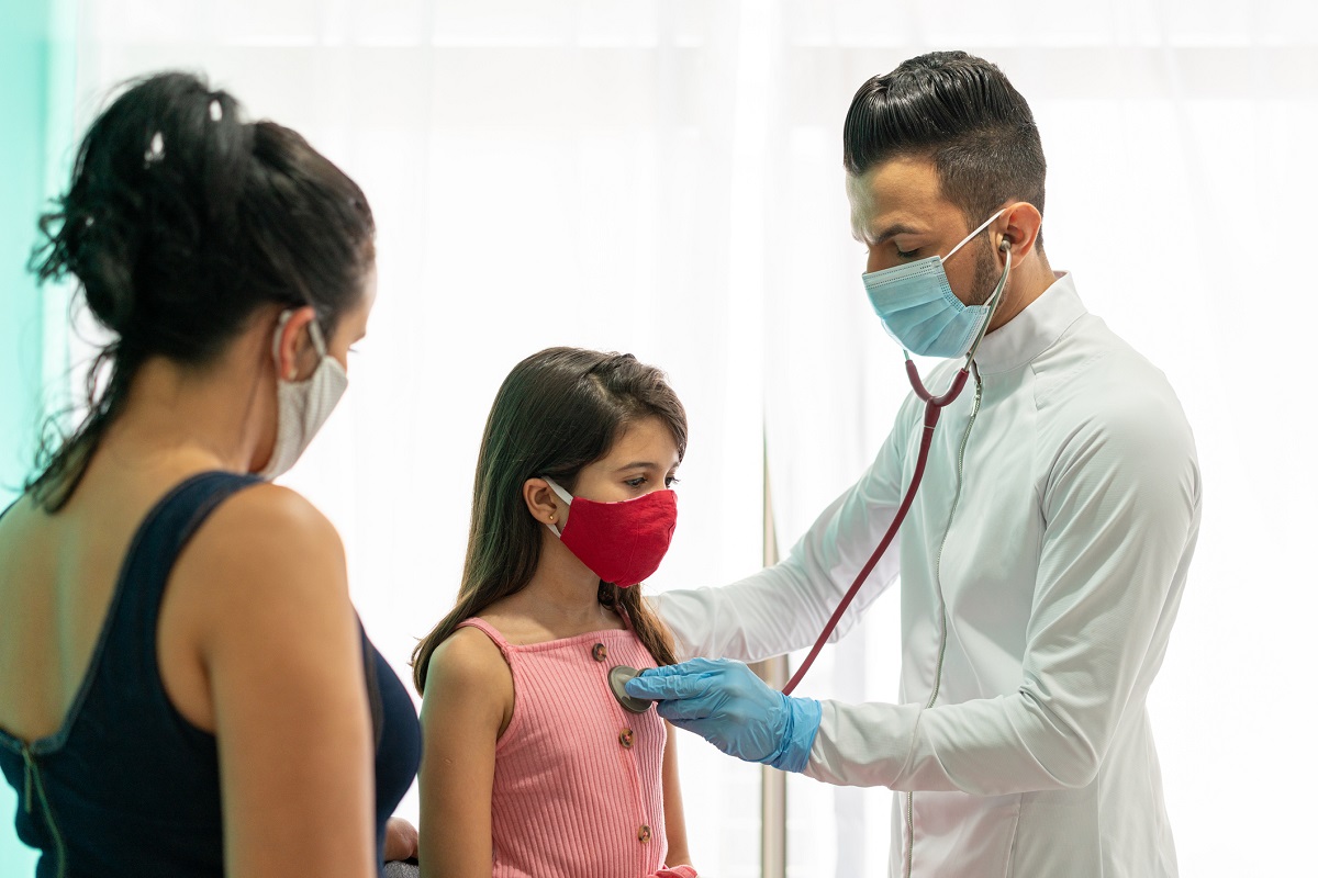 A doctor holds a stethoscope to the heart of a young girl as a woman looks on.