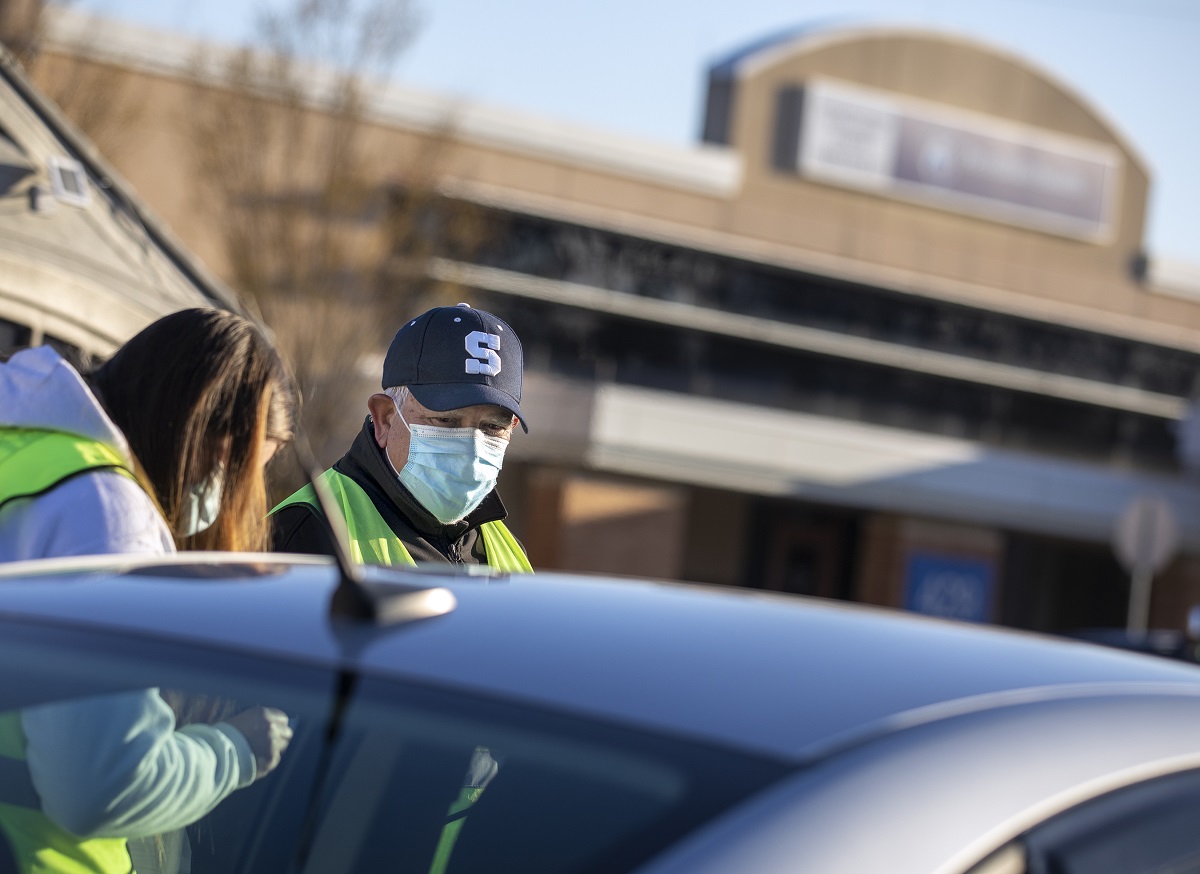 A man in a ball cap with an S on the front and a surgical mask stands next to a woman in a mask and a hoodie, partially obscured by a car.