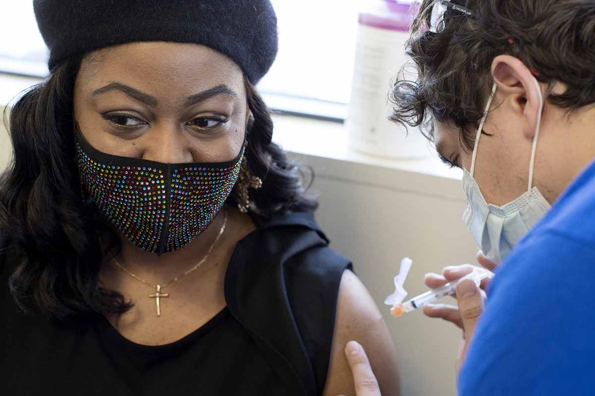 Tamika Washington, que lleva una máscara facial con joyas, un collar de crucifijo y una boina, mira lejos de su brazo desnudo mientras un trabajador de salud se prepara para insertar una jeringa llena con la vacuna COVID-19.