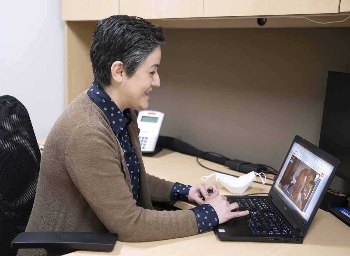 Una médica con suéter y camisa de vestir se sienta en una computadora y sonríe mientras habla con un paciente durante una visita de tele salud.