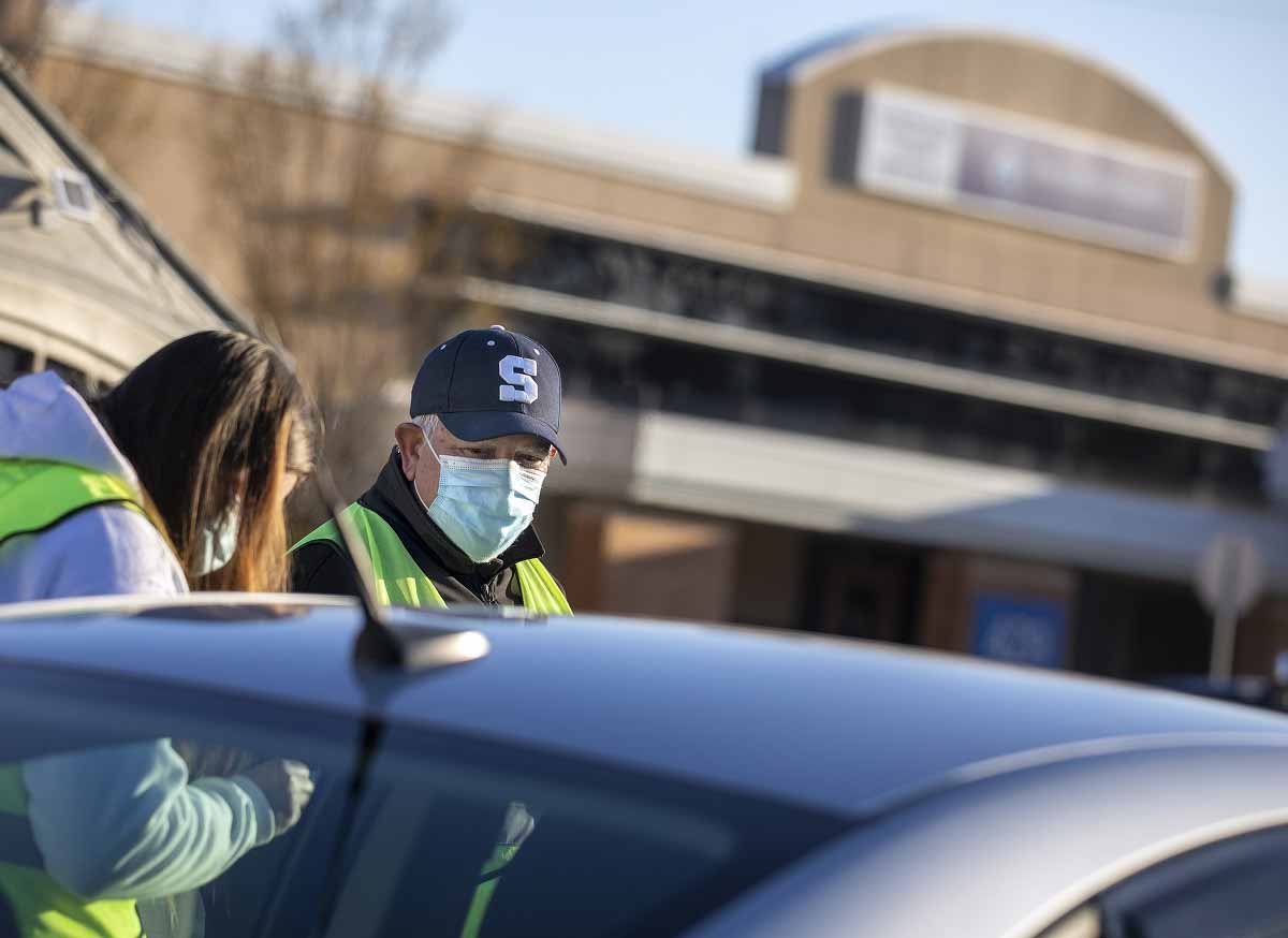 Un hombre con una gorra de béisbol con una S en la parte delantera y una máscara quirúrgica se para junto a una mujer con una máscara y una sudadera con capucha, parcialmente oscurecida por un auto.