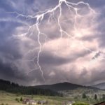 A thunderstorm and lightning flashes in the sky over hills with several houses.