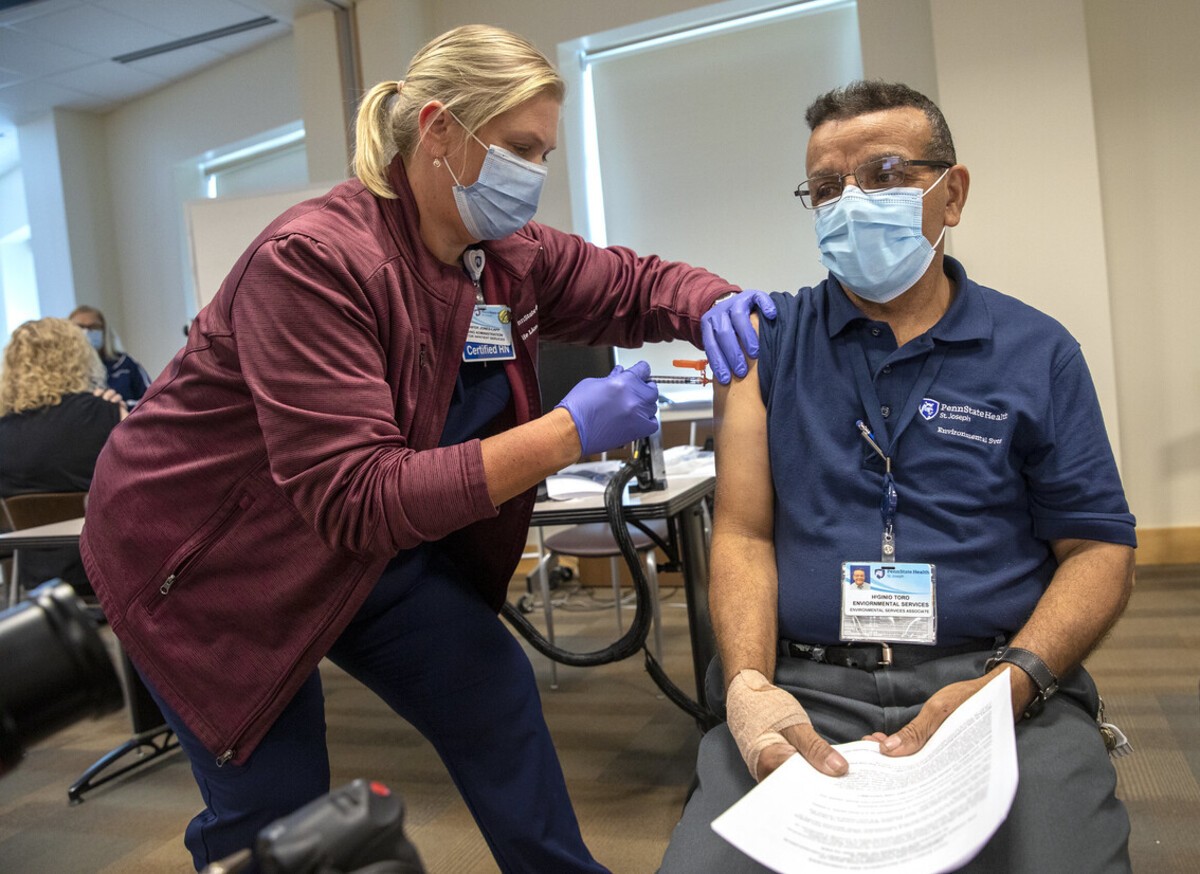 A woman wearing a lab coat, face mask and gloves vaccinates a man wearing a face mask and glasses.