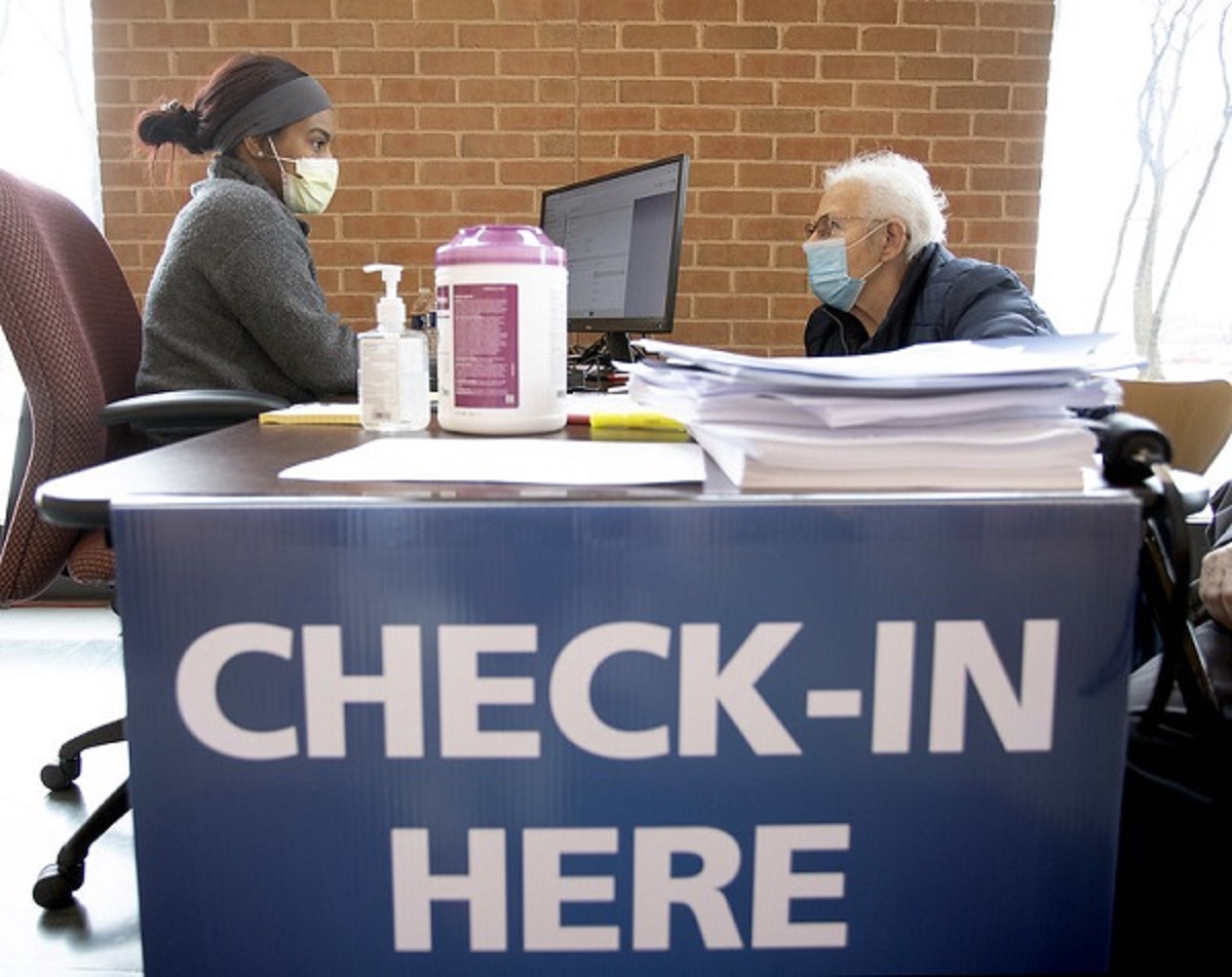 A woman with her hair pulled back and wearing a head band and face mask sits at a table in front of a computer facing an elderly woman wearing a face mask and glasses A sign on the table says, “Check-in here.”