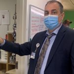Man in suit holding tape measure points to a silver sticker on the doorframe of a room in a hospital.