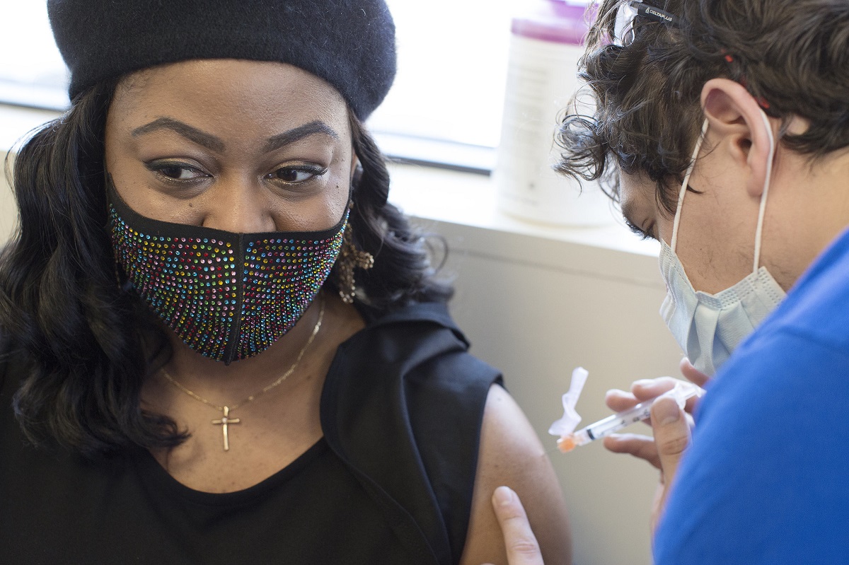 A woman in a decorated surgical mask and a beret receives an injection.