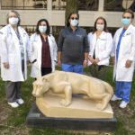 A group of male and female kidney and liver transplant team members, wearing masks, stand side-by-side behind a statue of the Penn State Nittany Lion.
