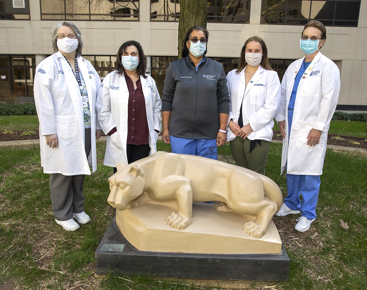 A group of male and female kidney and liver transplant team members, wearing masks, stand side-by-side behind a statue of the Penn State Nittany Lion.