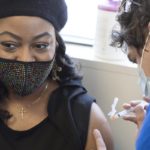 Tamika Washington, who wears a jeweled face mask, a cross necklace and a beret, looks away from her bare arm as a health care worker prepares to insert a syringe filled with the COVID-19 vaccine.