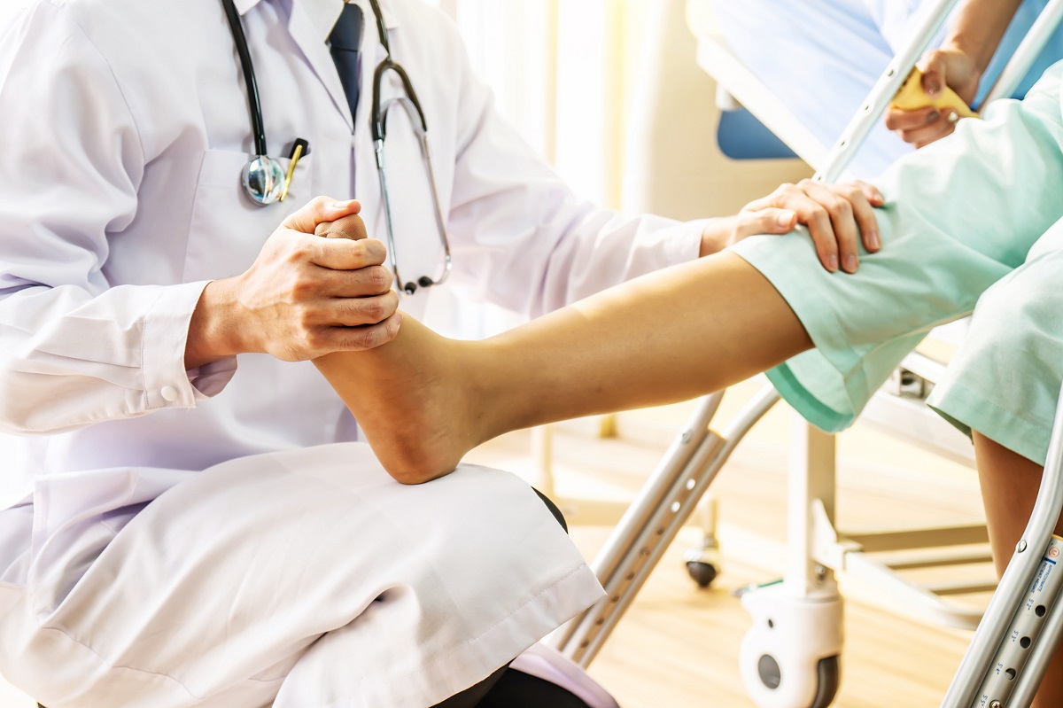 A doctor touches a patient’s foot and knee during an exam. The doctor is wearing a white coat and a stethoscope. The patient is wearing short pants. Their faces are not visible.