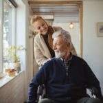 A woman pushes an older man in a wheelchair, as both smile. They are next to a sunny window.