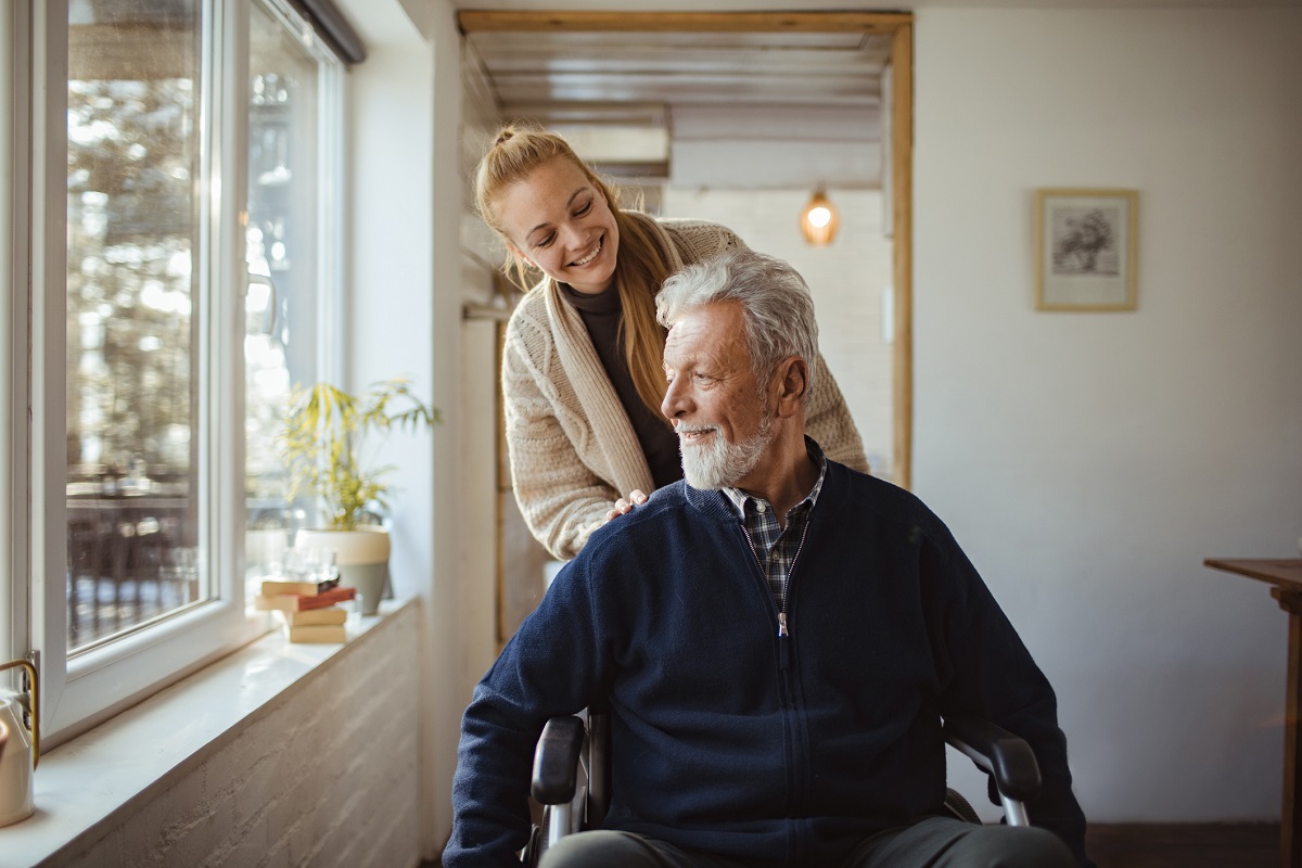 A woman pushes an older man in a wheelchair, as both smile. They are next to a sunny window.