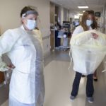Two women don personal protective equipment in a hospital hallway.