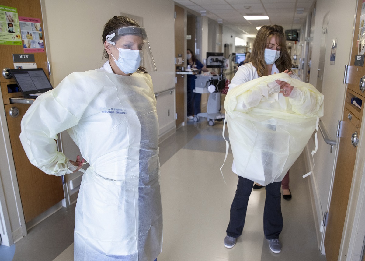 Two women don personal protective equipment in a hospital hallway.