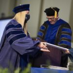 A woman in a graduation cap and gown receives an envelope from someone off camera. Behind her, a man at a lectern in a cap and gown smiles downward.