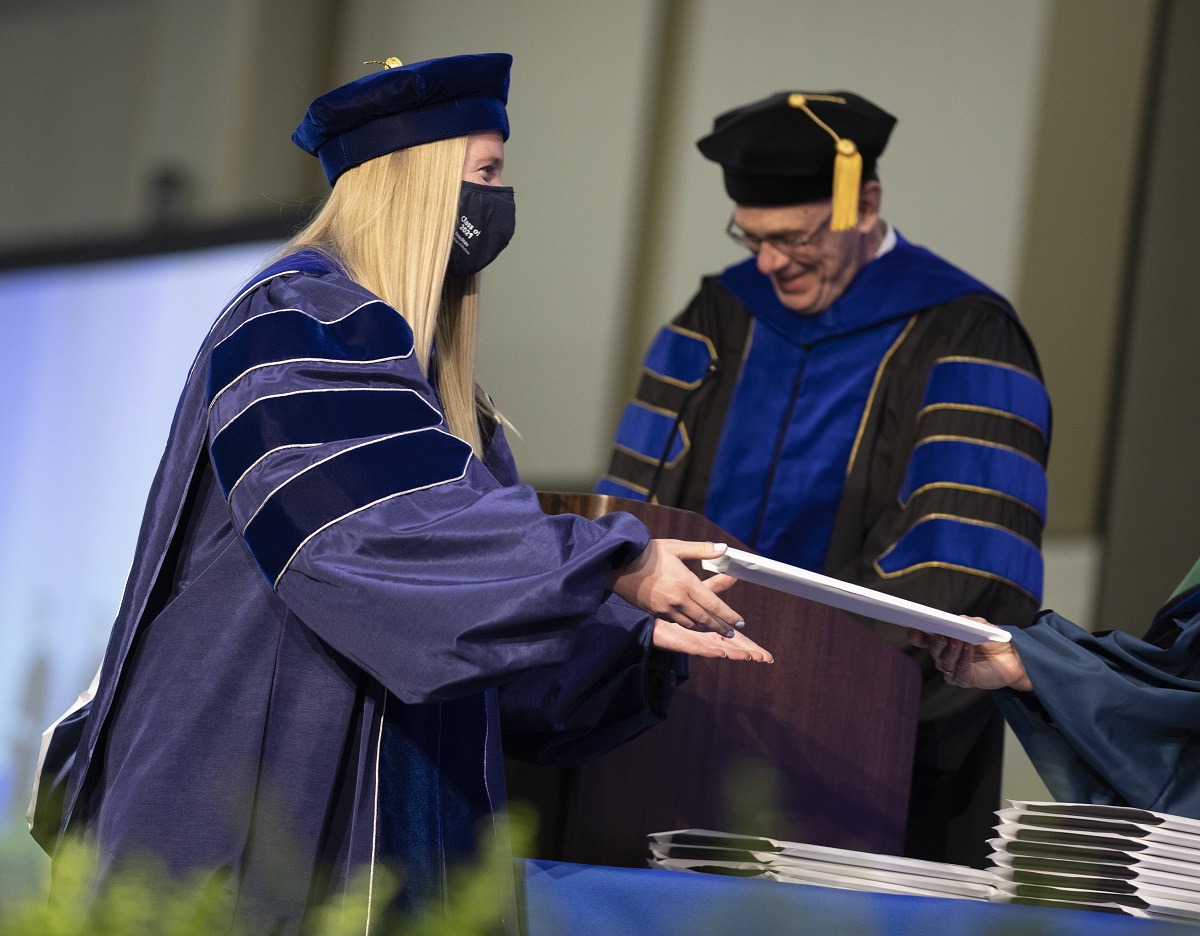 A woman in a graduation cap and gown receives an envelope from someone off camera. Behind her, a man at a lectern in a cap and gown smiles downward.