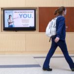 A woman with a backpack walks past a TV screen on a wooden wall.