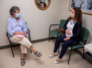 A female medical care provider, seated on the left, chats with a pregnant patient seated on the right.