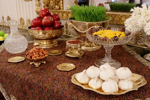This photo from Wikimedia COmmons shows a traditional Nowruz table arrangement. Platters and dishes filled with nuts, apples and other food are arranged on a brocade tablecloth. A fancy mirror hangs in the background.