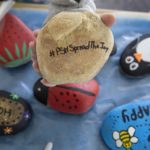 Painted rocks featuring messages such as “bee happy” and “hope” sit on a table. Other rocks are painted to resemble a penguin, a lady bug and a strawberry. A tan rock that reads “#PSHSpreadTheJoy” sits in the palm of a hand shown above the other rocks.