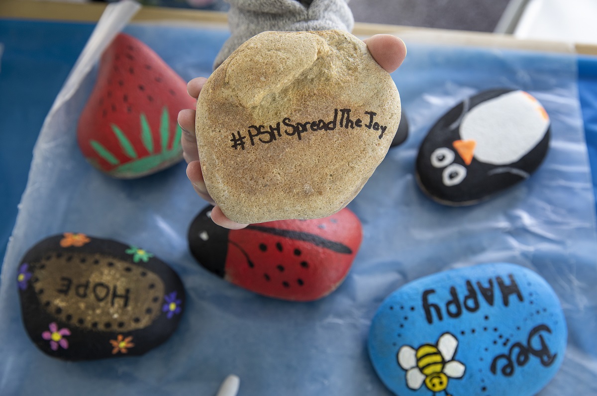 Painted rocks featuring messages such as “bee happy” and “hope” sit on a table. Other rocks are painted to resemble a penguin, a lady bug and a strawberry. A tan rock that reads “#PSHSpreadTheJoy” sits in the palm of a hand shown above the other rocks.