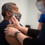 A man wearing a mask gets the COVID-19 vaccination in his shoulder. His eyes are closed.