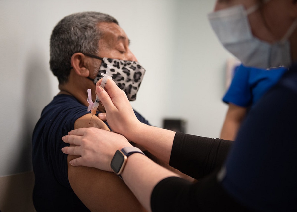 A man wearing a mask gets the COVID-19 vaccination in his shoulder. His eyes are closed.