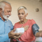 A woman has a blood pressure cuff on her left arm. A man standing next to her holds the monitor for it.