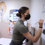 A woman in a surgical mask applies a paintbrush to a hospital room.