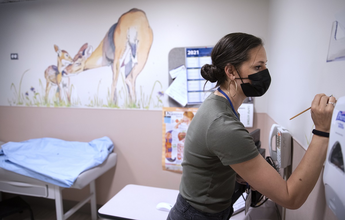 A woman in a surgical mask applies a paintbrush to a hospital room.