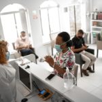 A woman wearing a flowered top and face mask speaks to a medical office professional wearing a white medical jacket and face mask at a reception desk in a medical outpatient lobby.