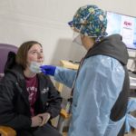 Michelle Mentzer stands over Holly Bressler and swabs her nose to test for COVID-19. Mentzer is dressed in personal protective equipment and wears glasses. Bressler is seated in a chair, hands clasped and looking upward. A computer sits on a cart in the background.