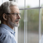 An older man wearing glasses looks out of a window.