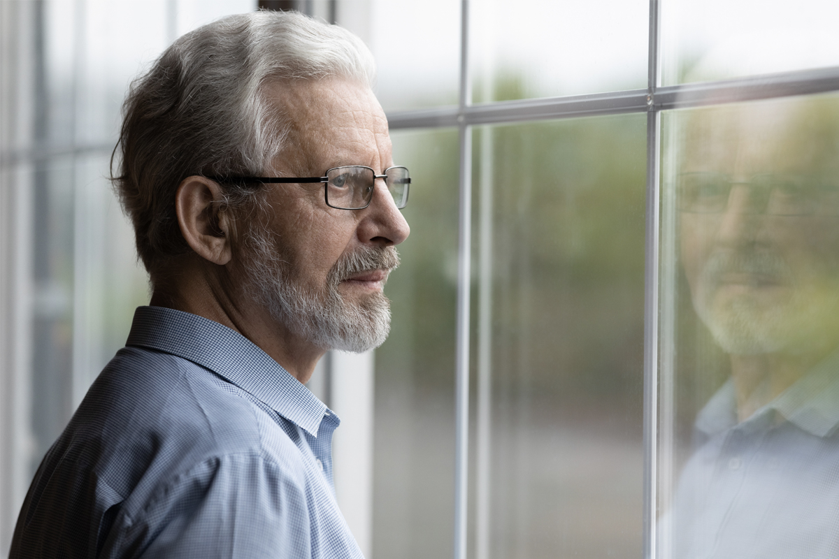 An older man wearing glasses looks out of a window.