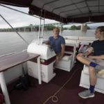 George Yatsky, who has short hair and wears glasses, shorts and a T-shirt, sits at the wheel of a pontoon boat and looks over at his son, who has a pony tail and wears a T-shirt, shorts and sneakers. A bridge stretched over a river in the background.