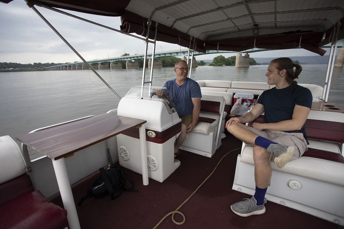 George Yatsky, who has short hair and wears glasses, shorts and a T-shirt, sits at the wheel of a pontoon boat and looks over at his son, who has a pony tail and wears a T-shirt, shorts and sneakers. A bridge stretched over a river in the background.