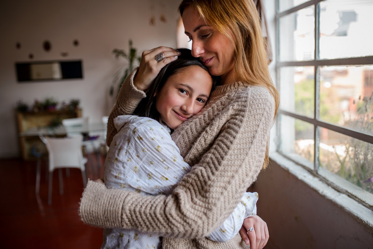 A girl smiles at the camera as she hugs her mother. The girl is wearing a long-sleeved top and has long hair. Her mother is wearing a sweater and has her hand on her daughter’s head. Behind them is a window.
