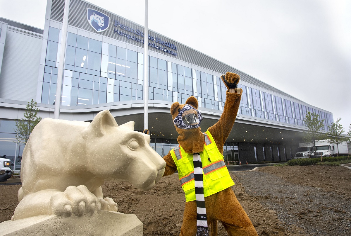 The Nittany Lion mascot raises its left arm as it touches the Nittany Lion statue with its left arm. The mascot is wearing a face mask, safety vest and scarf. Behind them is the newly constructed Penn State Health Hampden Medical Center, which has three stories and a sign that reads Penn State Health Hampden Medical Center. The top two stories are all glass windows.