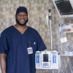 Nurse Viniquinn Terry, who has a beard and wears scrubs, smiles as he stands with medical equipment used in the cardiovascular intensive care unit.