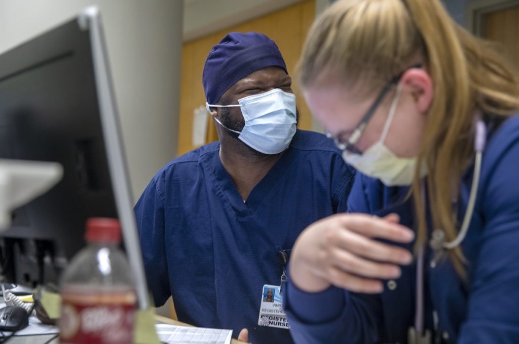 Viniquinn Terry, left, laughs with Amanda Sheets at Holy Spirit Hospital. Both are wearing scrubs and face masks. Viniquinn has a surgical cap on his head and a name badge. Amanda is looking down and leaning on her right arm. She has long, straight hair and is wearing glasses.